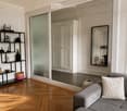 Modern living room with wooden herringbone flooring, grey sofa with decorative pillow, black shelving unit with various decorative items, large mirror, and a glass door leading to another room.