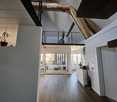 Open, modern hallway with wooden flooring, white walls, and a glass door leading to a living area. Features a metal railing and a small plant on a shelf.