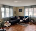 Modern living room with wooden flooring, large windows with grey curtains, a dark grey sectional sofa with multiple cushions, a small black coffee table, a white side table with a plant, and a bookshelf filled with books.