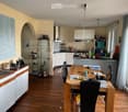 Bright kitchen with wooden flooring, white cabinets, and a central dining table with chairs. Includes a wine rack, sink, and various kitchen items on the counter.