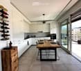 Modern kitchen with white cabinets, a wooden dining table, black chairs, and a wine rack on the wall. The space features large windows with a view of the balcony and outdoor area.