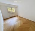 Empty room with wooden herringbone flooring, white walls, and a large window with a view of greenery. A radiator is mounted below the window.