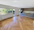Bright, unfurnished room with wooden herringbone flooring, large windows, and a modern kitchen area with grey cabinets and stainless steel appliances.