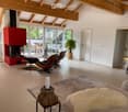 Bright living room with wooden floors, exposed wooden beams, large windows, fireplace, sofa, coffee table, ottoman, chairs, and a plant in the corner.