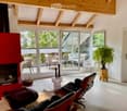 Bright living room with wooden ceiling beams, large glass doors leading to a patio, a red fireplace, and modern seating including a sofa and chairs.