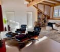 Modern living room with exposed wooden beams, white walls, and large windows. Features a cream sofa, black leather chairs, ottomans, a bookshelf, and a table lamp.