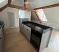 Modern kitchen with light wooden flooring, grey cabinets, black countertop, and built-in oven. Exposed wooden beams on the ceiling and large windows provide natural light.