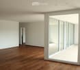 Empty room with wooden flooring, white walls, large glass sliding doors, and a visible adjacent room through an open doorway.