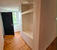Modern hallway with wooden flooring, built-in white shelving unit, and a black door leading to a room with a window.