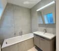 Modern bathroom with a bathtub, sink, and large mirror with built-in lighting. Grey tiles and sleek cabinetry.