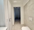 Modern bathroom with white tiles, a toilet, a sink, and a hallway leading to another room.
