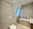 Modern bathroom with beige tiled walls and floor, featuring a toilet, bathtub, sink, and a skylight window providing natural light.