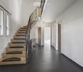 Modern hallway with black tiled floor, white walls, staircase with beige steps and black railings, several doors and windows.