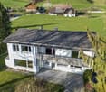 A modern two-story house with a flat roof and large windows, surrounded by green fields and trees. The house features a covered entrance and is situated in a rural landscape with additional buildings visible in the background.