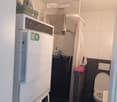 Compact bathroom with a washing machine and dishwasher stacked, a toilet, a shower area with a curtain, and cleaning supplies on a shelf.