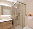 Modern bathroom with a glass-enclosed shower, wooden vanity with sink, mirror, and toilet. Features include a towel, candle, and decorative items on the shelf.