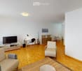 Bright living room with wooden flooring, white walls, and large windows. Features a beige sofa, coffee table, TV stand, floor lamp, and a small dining area with chairs and a table.