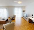 Modern living room with light wooden flooring, large windows with white curtains, a sofa, coffee table, armchairs, TV stand, and a desk with a chair.