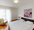 Modern bedroom with a large bed, nightstands, a wooden dresser, a chair, and a decorative vase on the dresser. The room features large windows with white curtains and a colorful abstract painting on the wall.