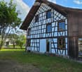 Traditional half-timbered house with light blue walls and dark wooden beams, surrounded by a grassy yard and trees.