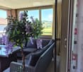 Balcony with a wicker sofa, glass table, and decorative plant with purple flowers, overlooking a scenic countryside view.