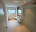 Modern bathroom with a toilet, double sink, mirror, and glass shower enclosure. Light-colored tiles and a window provide natural light.