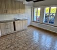 Empty kitchen with light wooden cabinets, granite countertop, sink, and large windows overlooking a parking area.