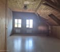 Empty attic space with wooden walls, ceiling beams, and large windows, featuring a smooth wooden floor and simple radiator heating.