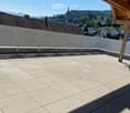 A spacious, sunlit balcony with a tiled floor and wooden beams, overlooking a scenic town with visible rooftops and distant mountains.