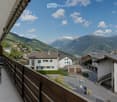 Balcony with wooden railing, overlooking a scenic mountain valley with houses and a road below.