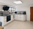 Modern kitchen with white cabinets, stainless steel appliances, and a wooden floor. Features include a refrigerator, oven, sink, coffee maker, and a small dining area with a chair and table.