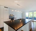 Modern kitchen with white cabinets, black countertop, stainless steel sink, and built-in oven. Large windows and sliding doors lead to a balcony.