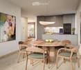 Modern dining area with a round wooden table and six chairs, adjacent to a sleek kitchen with grey cabinets, stainless steel appliances, and a pendant light. Artwork adorns the walls, and a fruit bowl sits on the table.