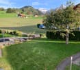 Well-maintained garden with green lawn, gravel pathways, small water feature, and surrounding trees. Scenic mountain view in the background.