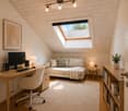 A cozy attic room with wooden floors, a skylight window, a white sofa, a wooden desk with a laptop, a bookshelf, and a small plant. The room features warm lighting and a textured rug.