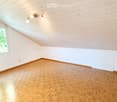 Empty room with wooden herringbone flooring, white walls, and exposed wooden ceiling beams. Features a single window and built-in lighting fixture.