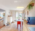 Open-plan kitchen and living area with modern furniture, including a round dining table with chairs, a coffee machine, a sofa, and a refrigerator. The space features light-colored walls, wooden flooring, and a hanging plant.