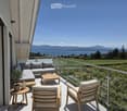 Balcony with a modern glass railing, overlooking a scenic lake and mountains. Features a grey sectional sofa, wooden coffee table, two wooden chairs, and a small potted plant.
