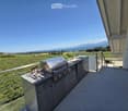 Balcony with a modern stainless steel grill, sink, and cutting board, overlooking a scenic countryside with mountains and a lake in the background.