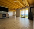 Open-plan living and dining area with exposed wooden beams, hardwood flooring, large windows offering a scenic view, and a modern oven and wood stove.