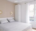 Bright bedroom with a white bed, grey pillows, a tall white wardrobe, and two decorative wall-mounted hat racks. The room features wooden flooring and large windows with white curtains, offering a view of a cityscape with balconies.