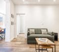 Modern living room with light wooden flooring, grey sofa, coffee table, dining area with chairs, and large windows with white curtains.