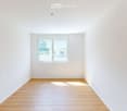 Empty room with light wooden flooring and white walls, featuring a large window with a view of greenery outside.