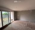 Empty room with unfinished walls and floor, large sliding glass door leading outside, visible construction materials and measurements on the left side.