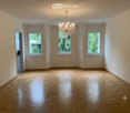Empty room with wooden parquet flooring, white walls, three windows, and a chandelier hanging from the ceiling.