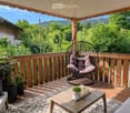Wooden balcony with a hanging wicker chair, small wooden coffee table, potted plants, and scenic countryside view.