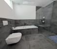 Modern bathroom with dark grey marble tiles, a white toilet, a bathtub, a glass-enclosed shower, and a towel on the floor.