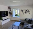 Modern living room with a grey sectional sofa, glass coffee table, flat-screen TV, built-in bookshelf, and potted plants.