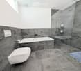Modern bathroom with grey marble tiles, a white toilet, a bathtub, a glass-enclosed shower, and a towel on the floor.