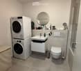 Modern bathroom with a white sink, mirror, toilet, washing machine, dryer, and a small cabinet under the sink.